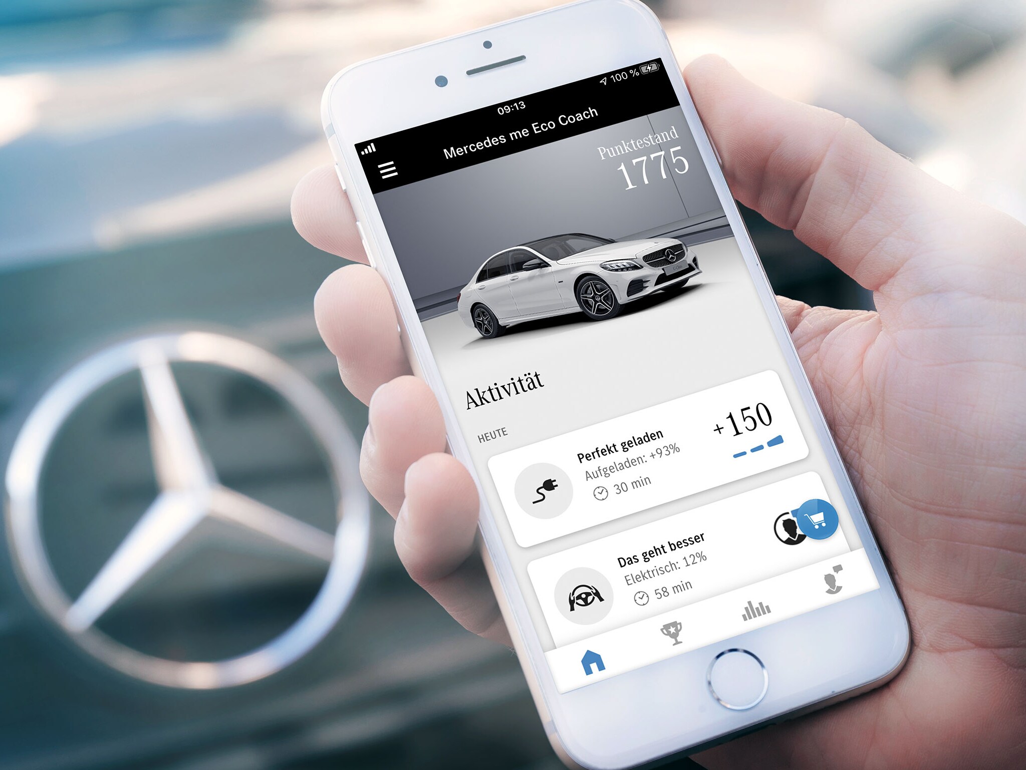 Egy Mercedes-Benz gépkocsi előtt egy személy a kezében egy okostelefont tart, amelyen meg van nyitva a Mercedes me Eco Coach alkalmazás