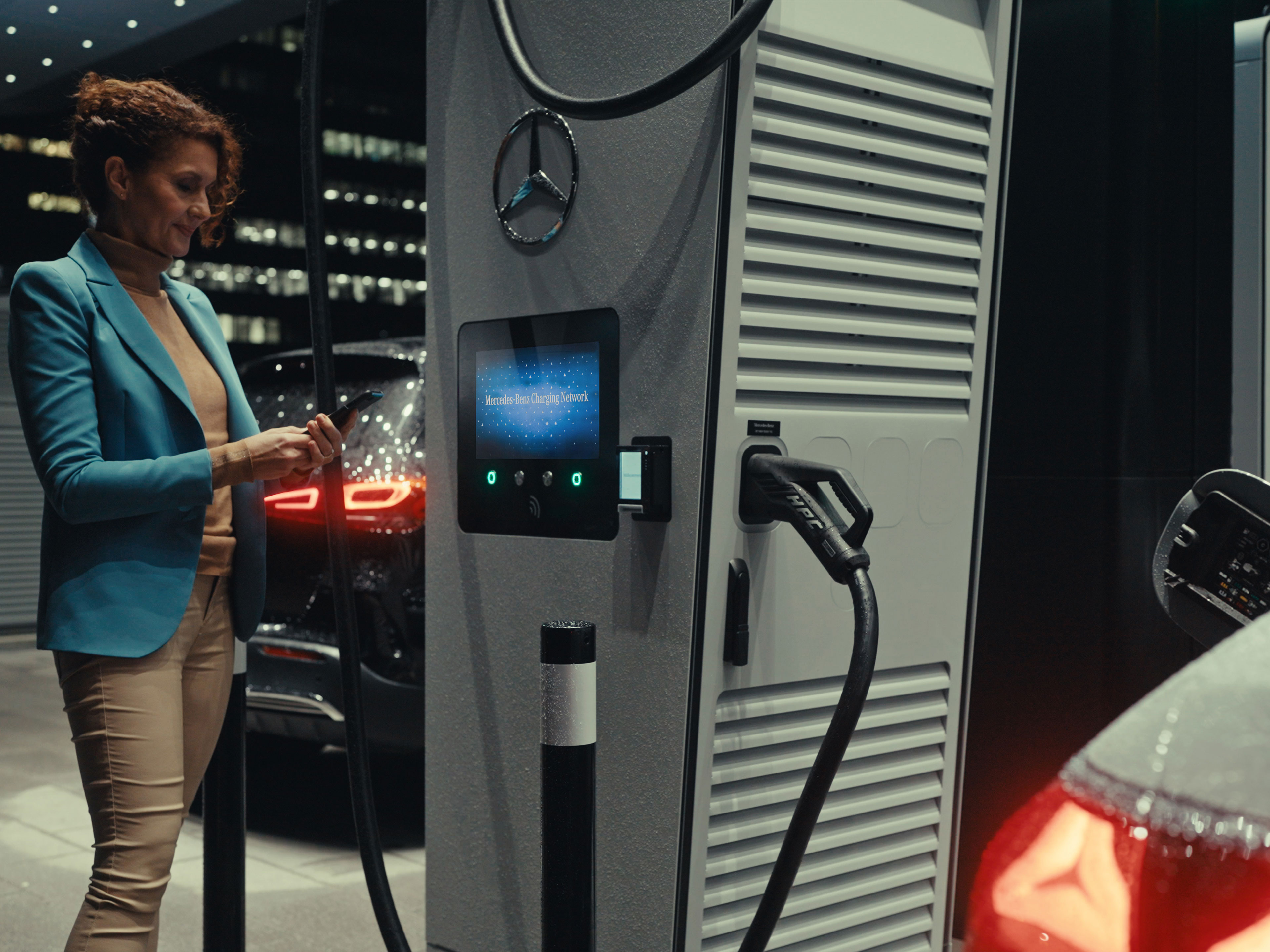 Mercedes-Benz alkalmazás | Töltés útközben | Mercedes-Benz Egy nő, kezében okostelefonnal, egy töltőállomás mellett áll.