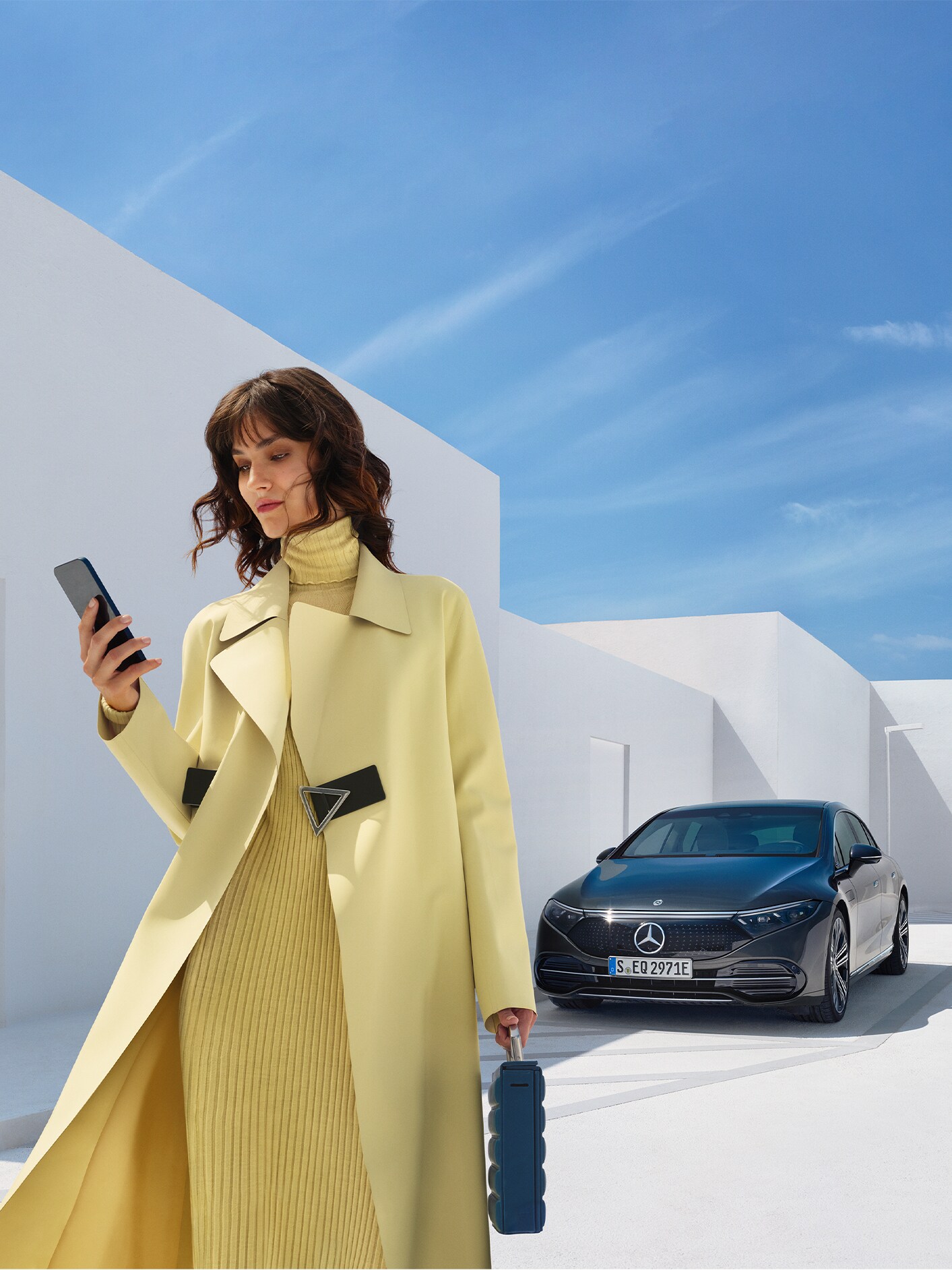 Egy hölgy ránéz a mobiltelefonjára, miközben elsétál elektromos Mercedes-Benz modelljétől.