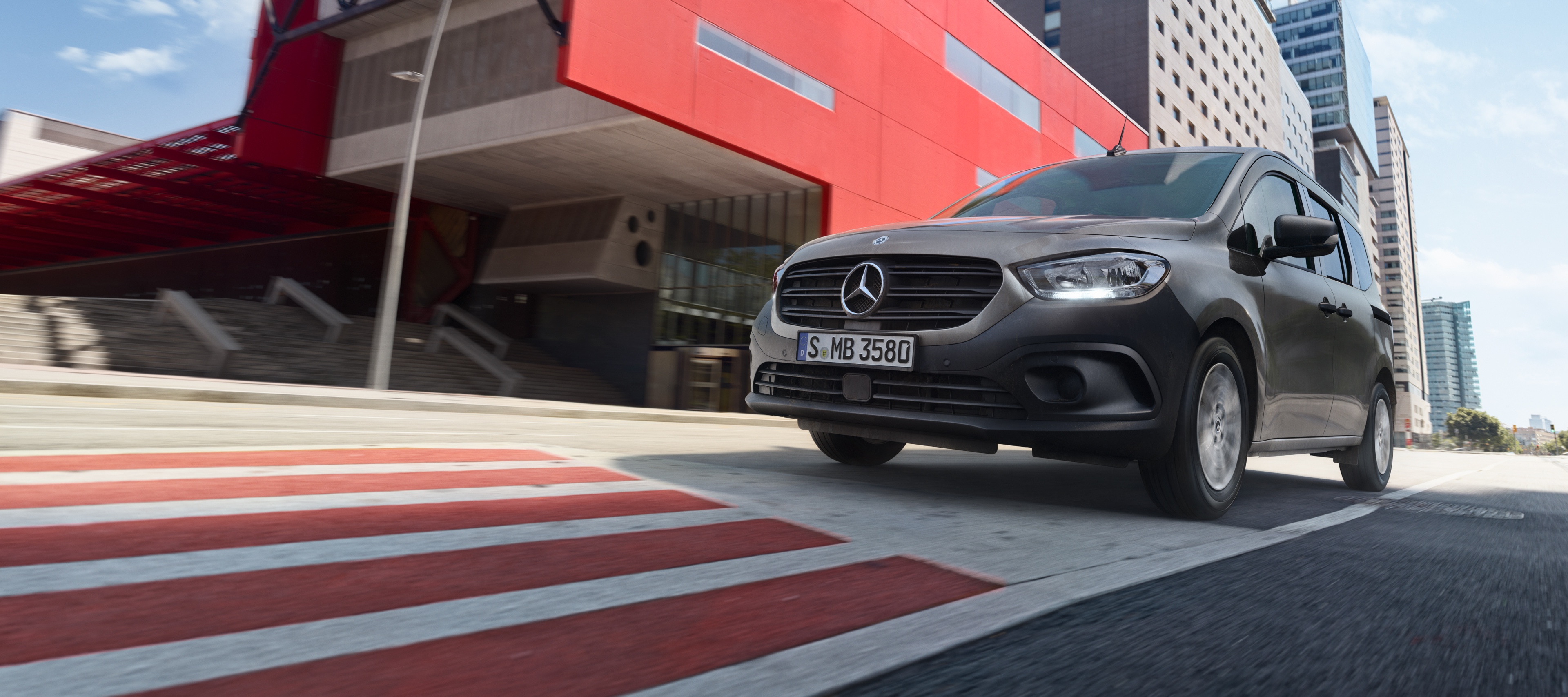 Citan Tourer | Mercedes-Benz kishaszongépjárművek A Citan Tourer, a Mercedes-Benz kompakt személyszállító furgonja egy modern városi területen halad keresztül.