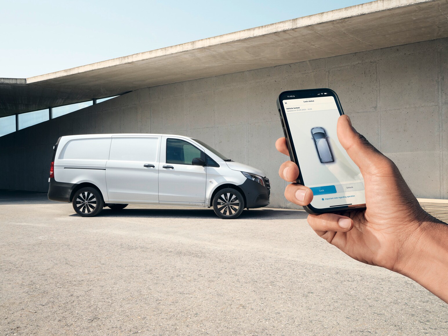 Egy okostelefont tartó kéz, amely a Mercedes me alkalmazást mutatja. A Vito zárt áruszállító a háttérben.