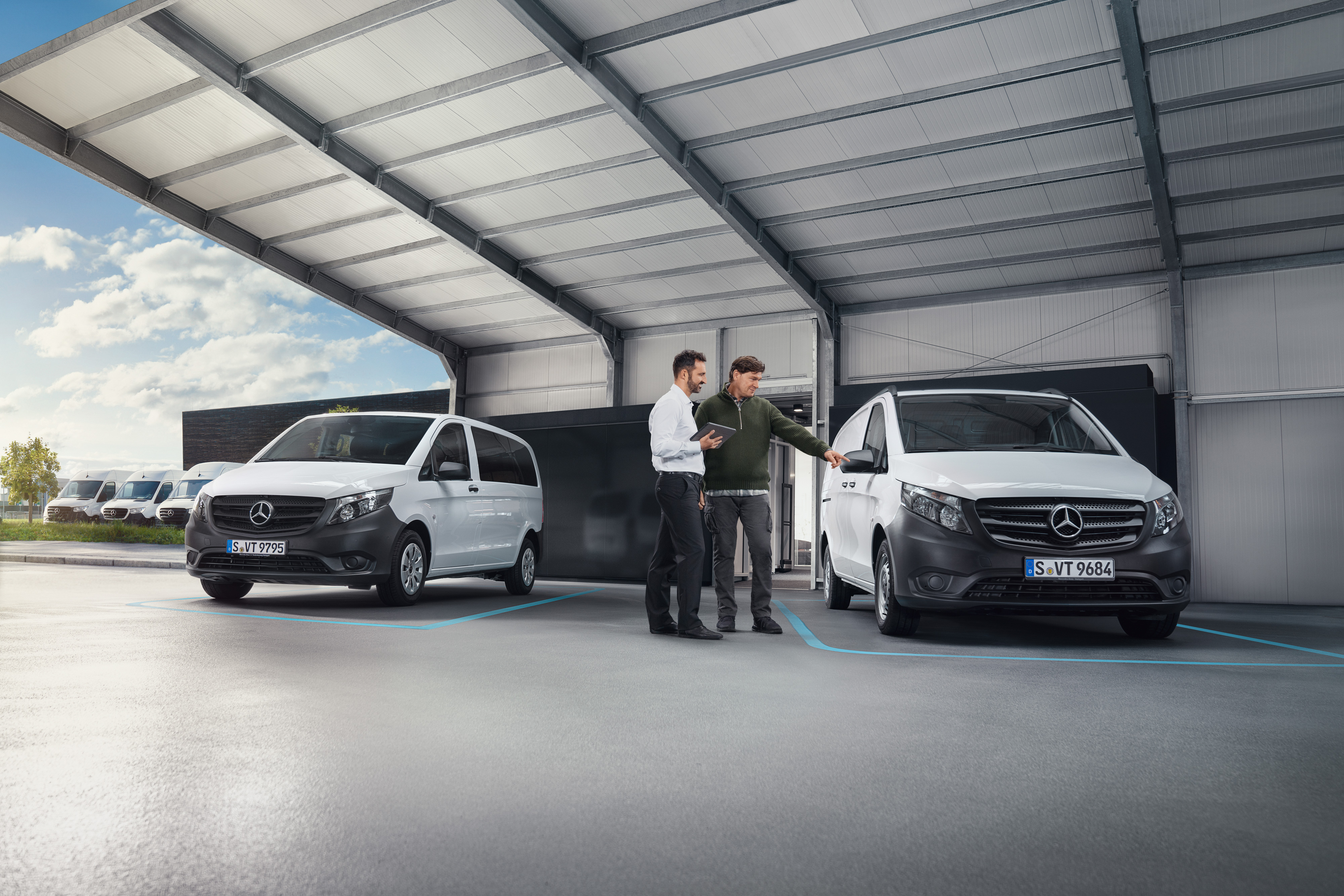 Mercedes-Benz | Komfort | Digitális ügyfélfogadás Két ember beszélget egy szervizben, két fehér Mercedes-Benz Vito előtt állva.
