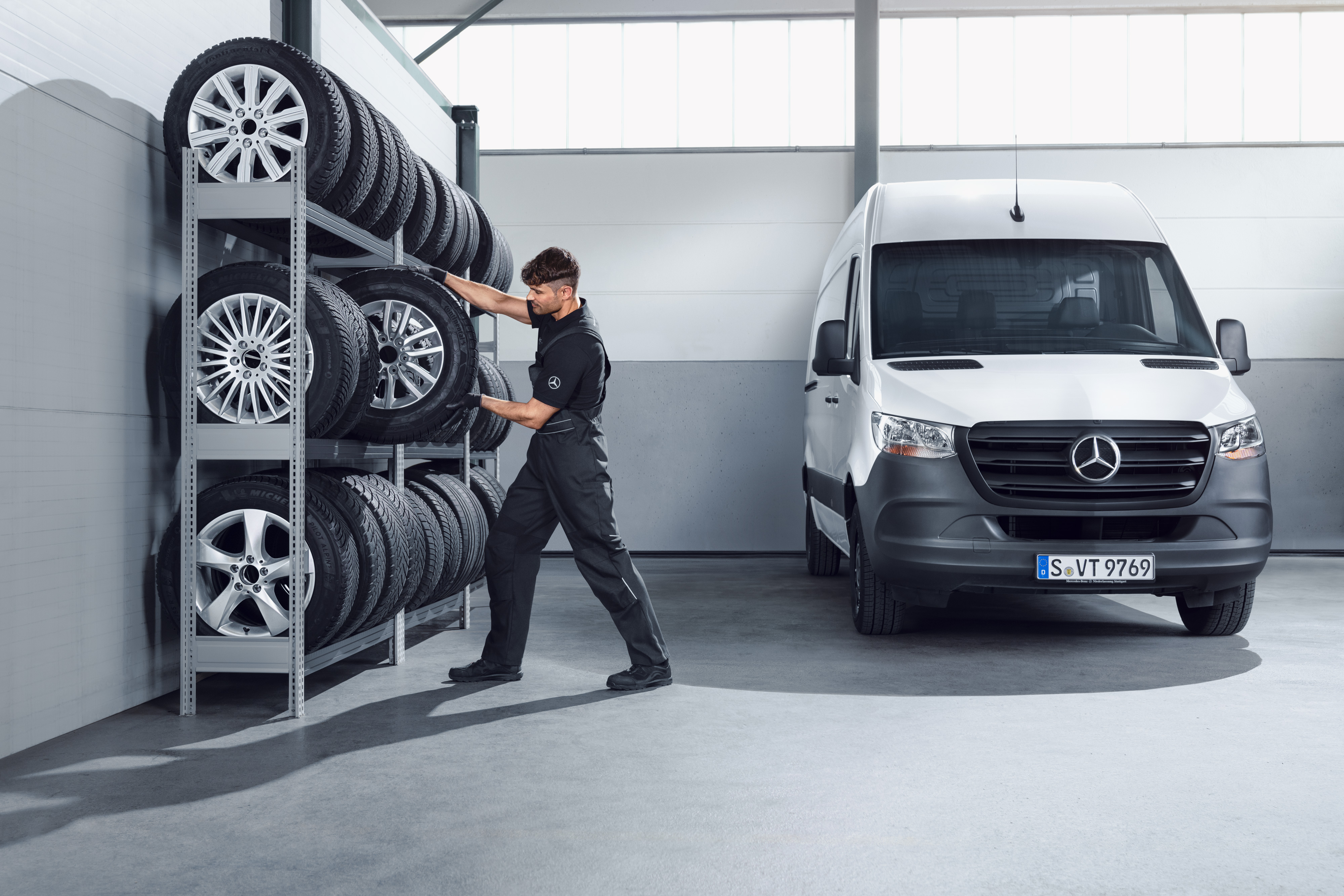 Mercedes-Benz | Kishaszongépjármű Eredeti Alkatrészek Egy szerelő kiválasztja a műhely polcáról a Mercedes-Benz komplett téli kerekeket egy fehér Sprinterhez.