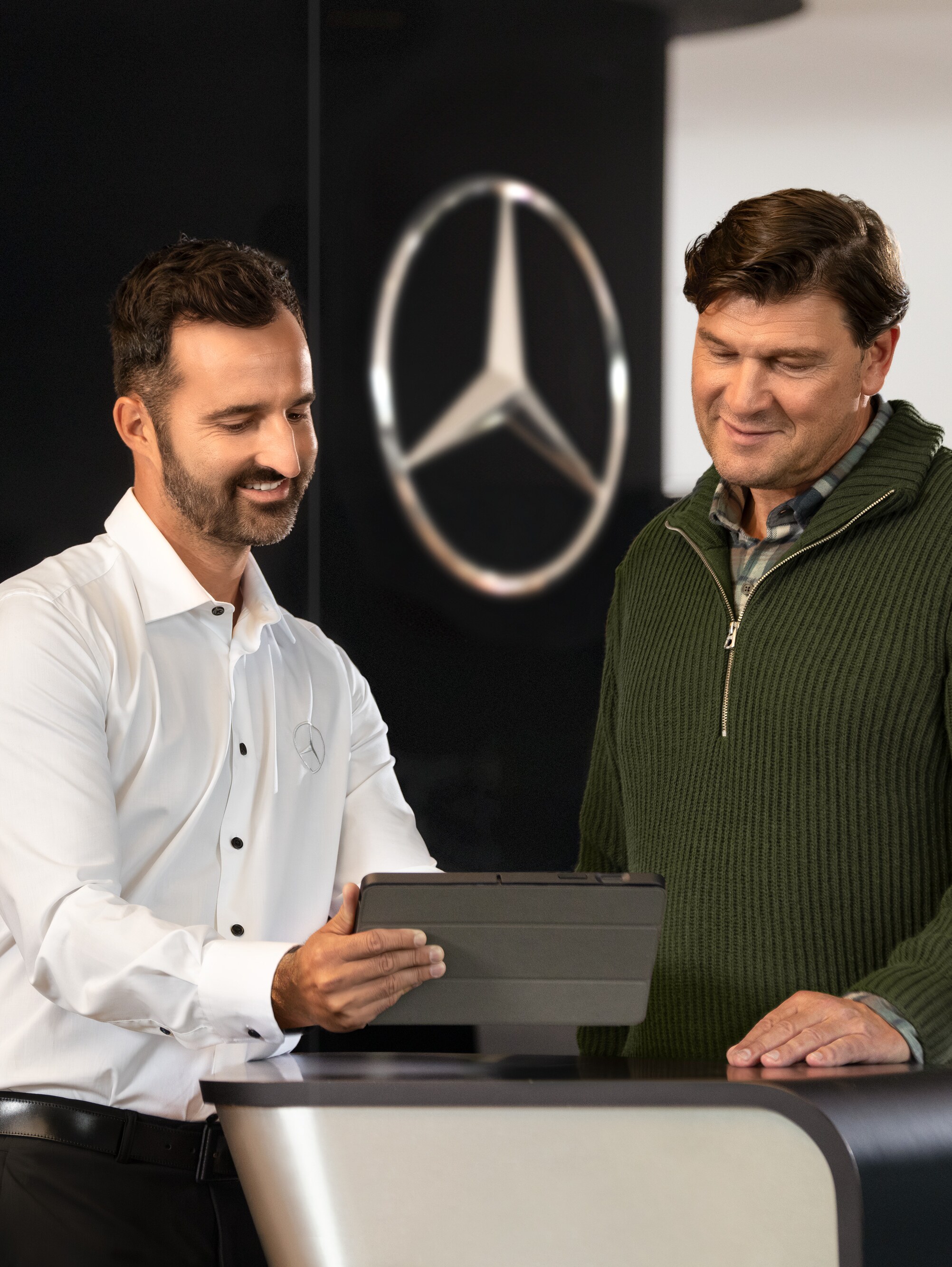 Két férfi beszélget egy pultnál, miközben egy tabletet néznek, a Mercedes-Benz csillag előtt.