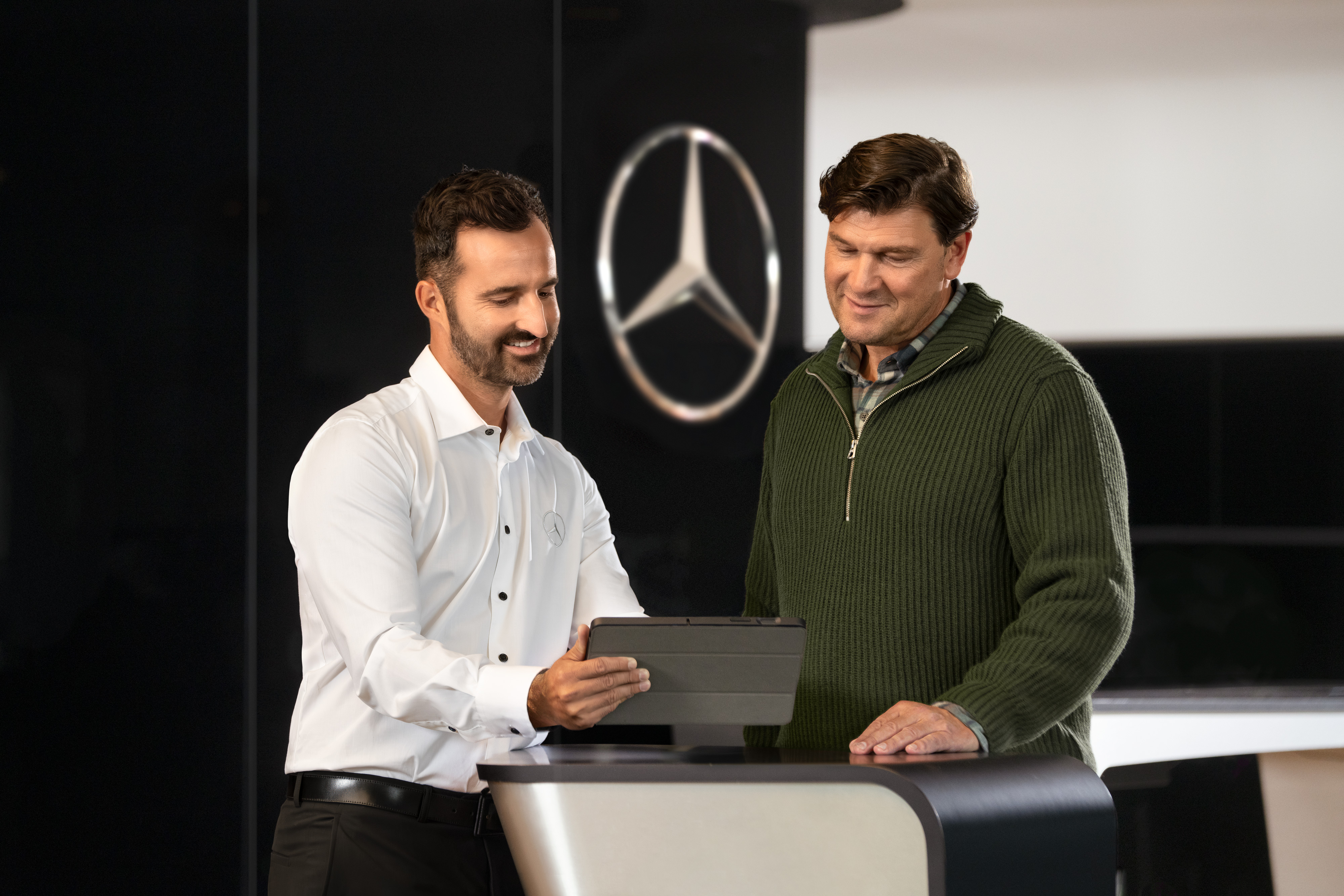 Mercedes-Benz | Szolgáltatások áttekintése Két férfi beszélget egy pultnál, miközben egy tabletet néznek, a Mercedes-Benz csillag előtt.