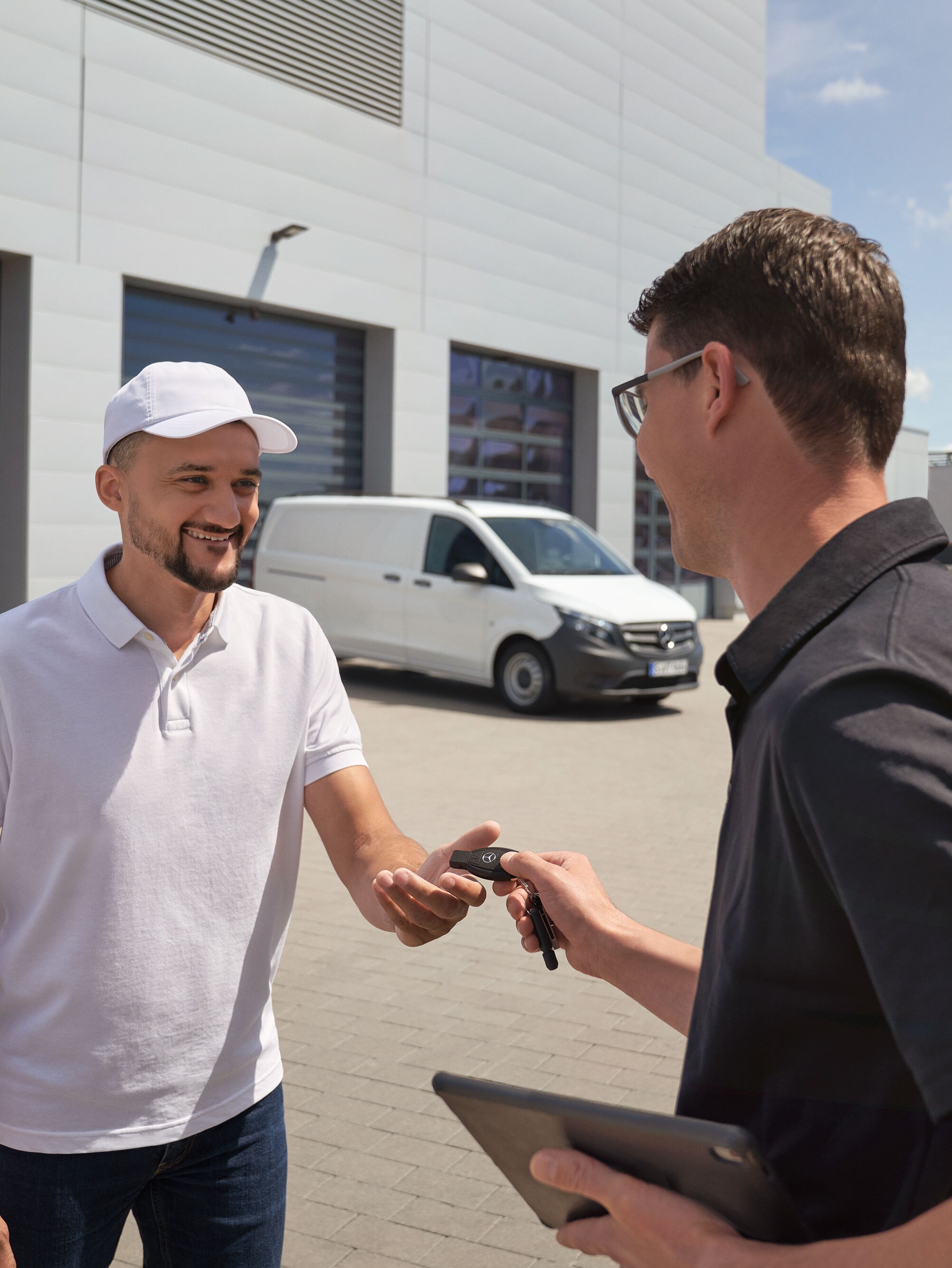 Egy szerelő átadja a kulcsot egy ügyfélnek, a háttérben egy fehér Mercedes-Benz Vito transzporter.