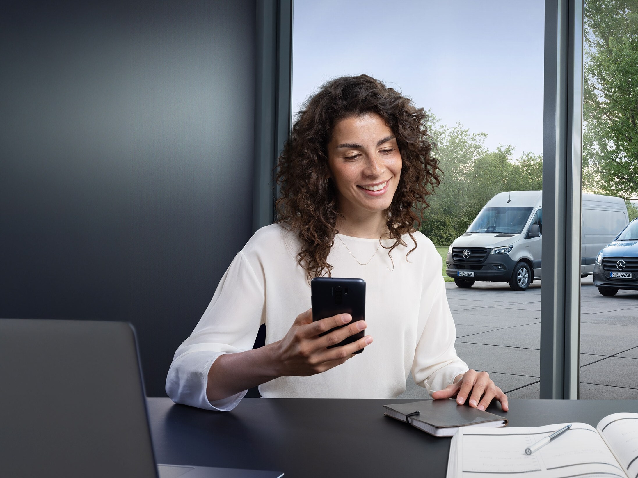 Egy nő okostelefonon keresztül vezérli több Mercedes-Benz transzporter flottafelügyeletét az iroda ablakában.
